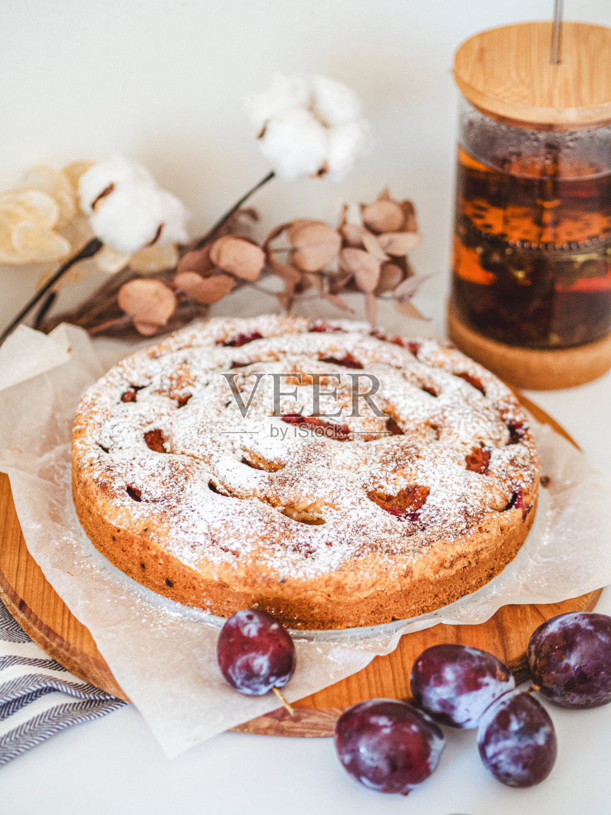 木板上放着糖粉和肉桂的李子派，旁边放着茶壶和干花。照片摄影图片