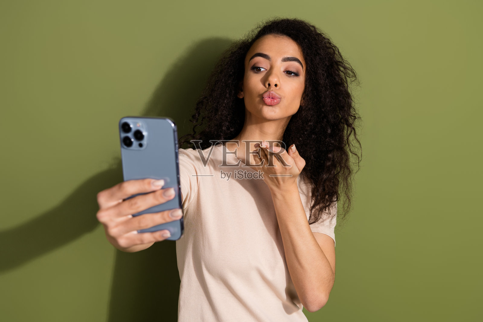 可爱的卷发女孩穿着米色t恤，自拍智能手机在卡其色背景下发送飞吻照片摄影图片