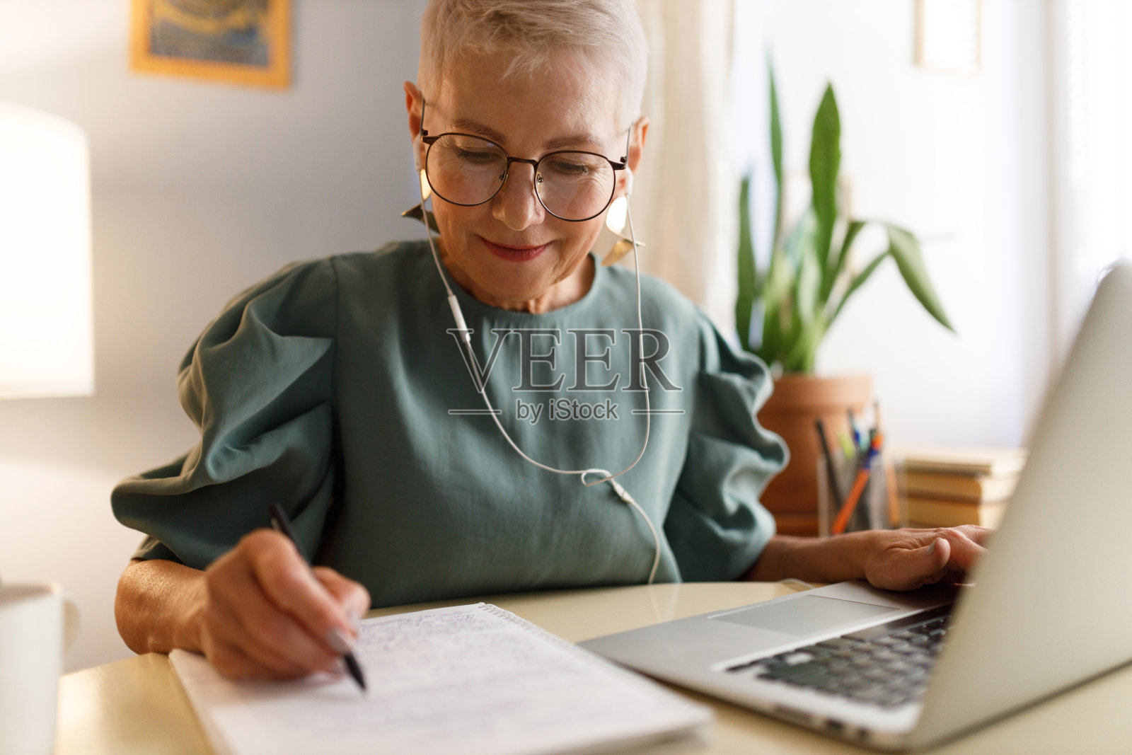 60多岁的优雅时尚女性，戴着连接笔记本电脑的白色有线耳机听在线课程或播客，坐在家里的桌子上，在课本上记下课程的主要内容照片摄影图片