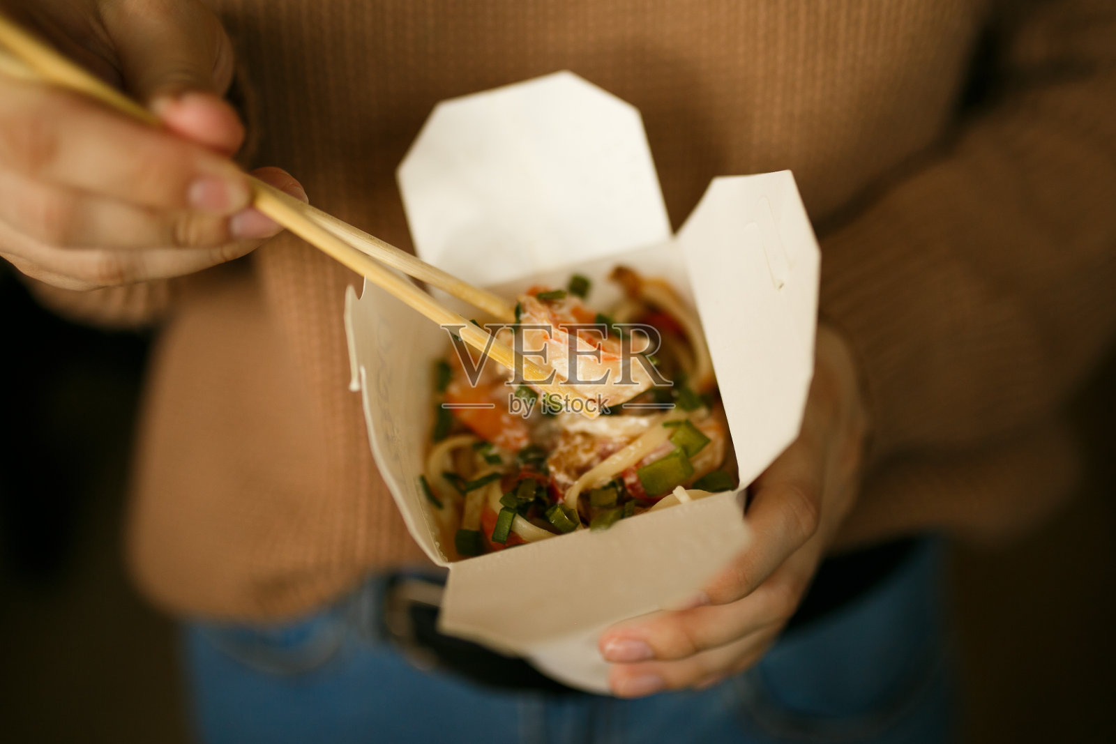 一个穿着便服的女孩坐在地板上吃着盒子里的面条，送餐照片摄影图片