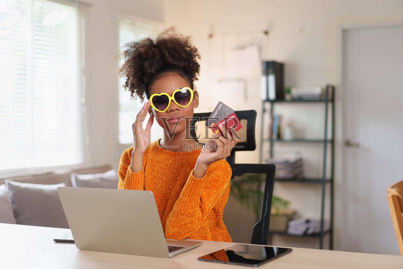 快乐的年轻非洲裔美国妇女戴着太阳镜，拿着信用卡，在舒适的家庭环境中对着笔记本电脑微笑。照片摄影图片
