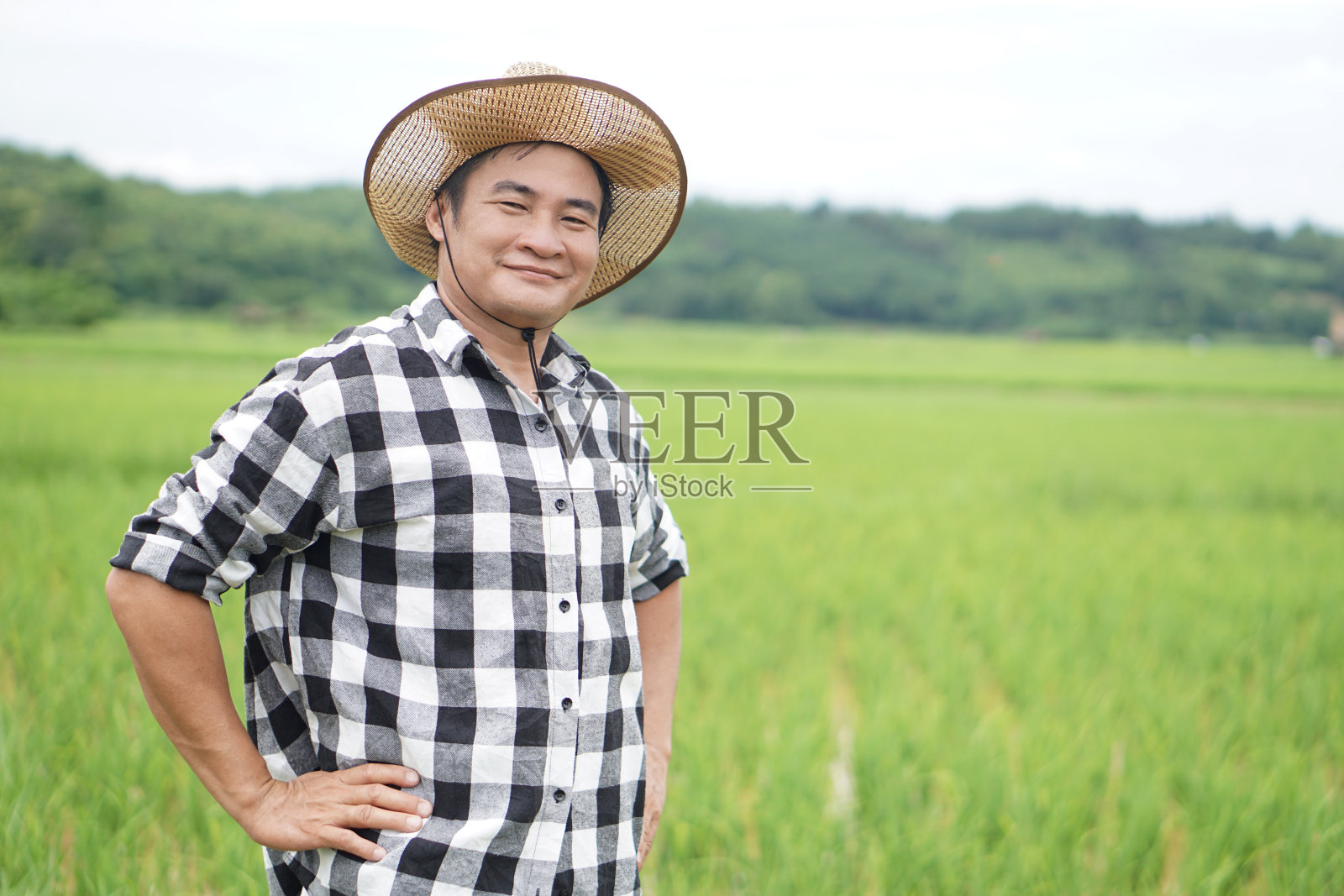 英俊的亚洲男子农民在稻田里，戴着帽子，穿着格子衬衫，双手放在腰上。自信。概念，农业职业。泰国农民。与大自然一起工作。有机农业照片摄影图片