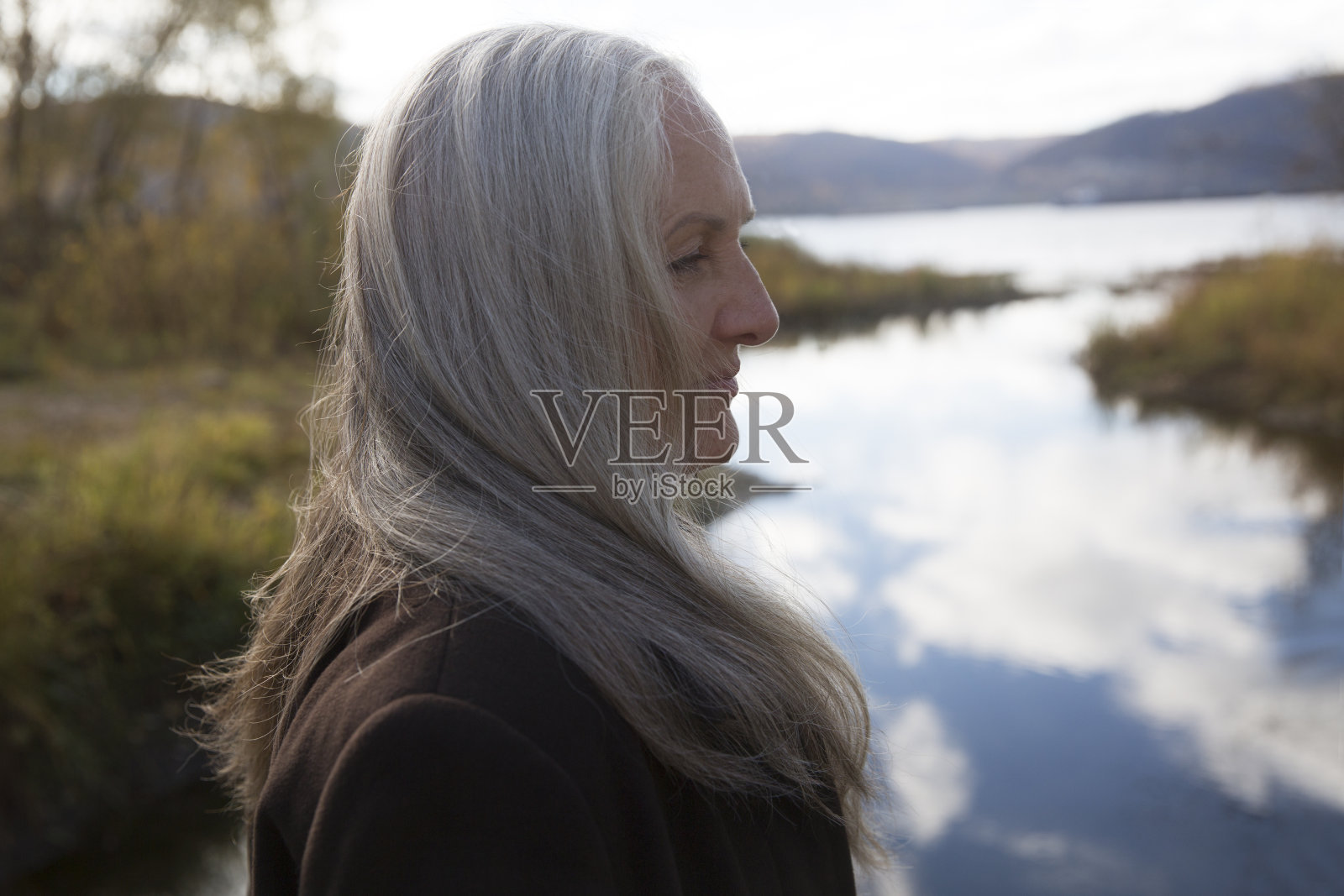 一位白发苍苍、身穿棕色外套的老妇人背靠着河岸站着。享受小事情。散步。世界精神卫生日。国际老年人日照片摄影图片