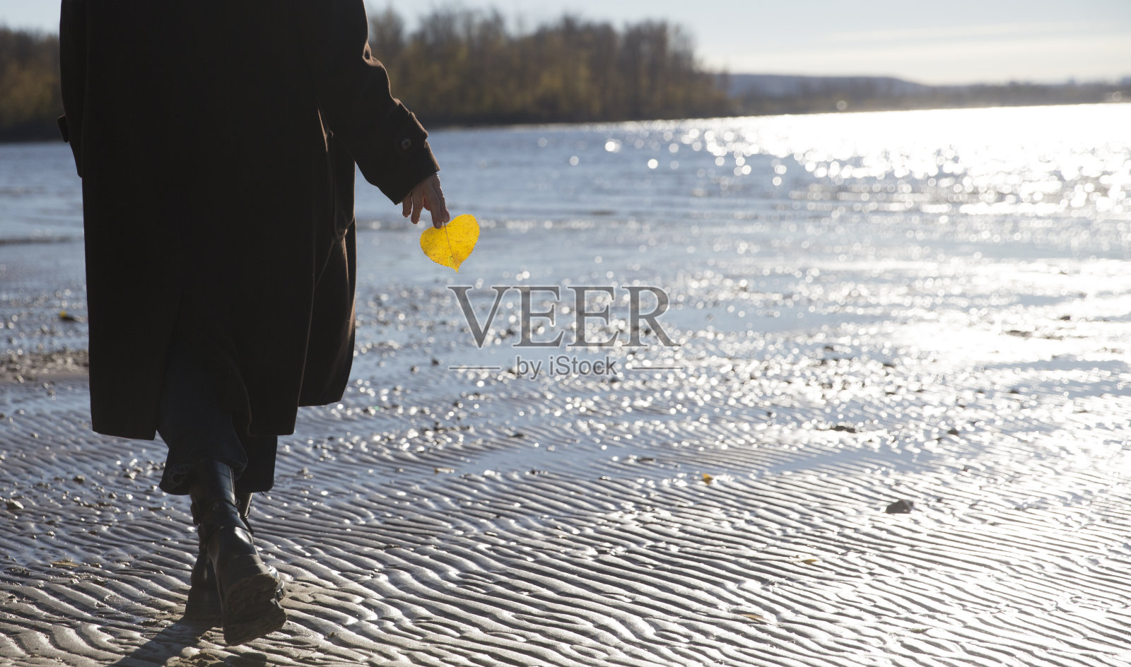 老妇人手持一片秋天的黄叶站在河边。节奏缓慢的生命。享受小事情。漫步在秋天的大自然中。照片摄影图片