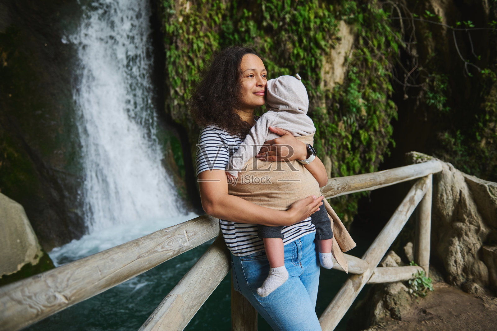 母亲抱着婴儿站在瀑布附近的木桥上，与家人一起户外自然漫步照片摄影图片