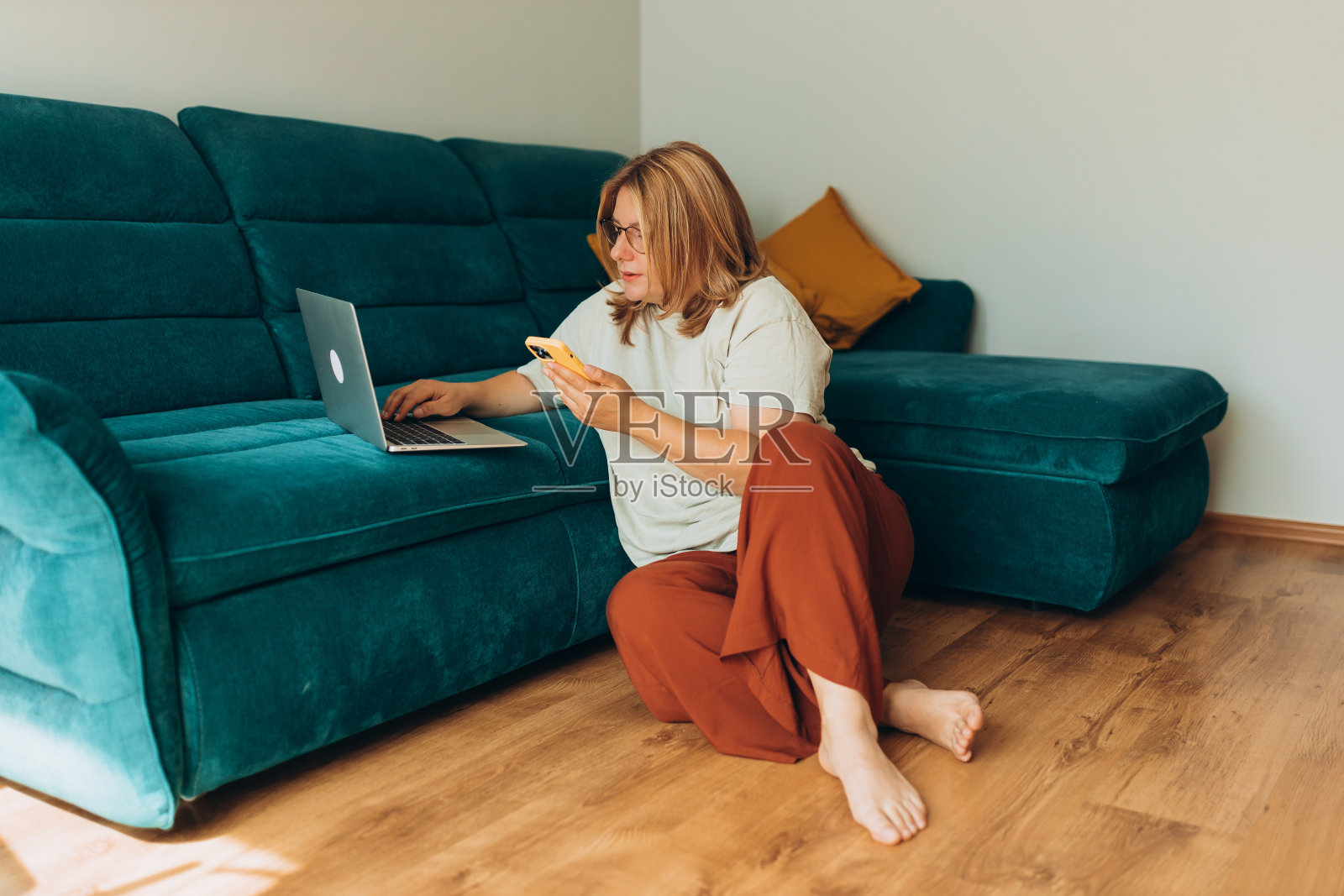 漂亮的女人戴着眼镜，拿着手机在家里沙发旁边的笔记本上工作。教育、工作和舒适的概念。30多岁的女性在室内工作。全身长度照片摄影图片