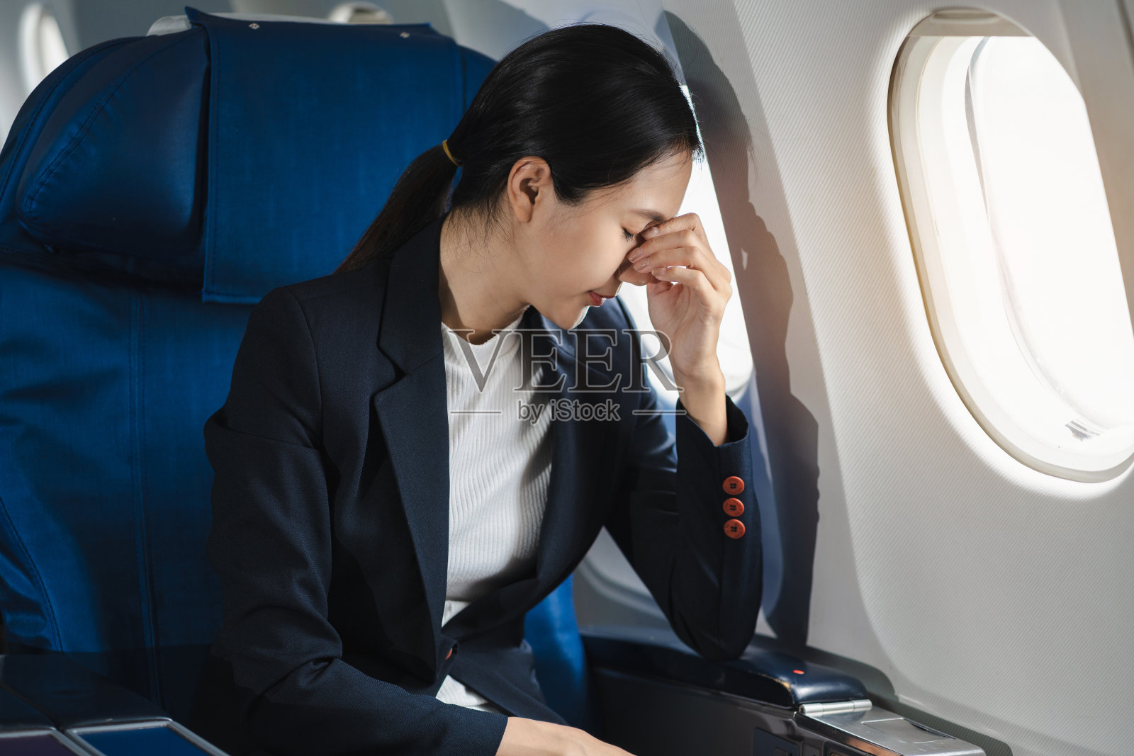 疲倦的亚洲妇女头痛感到恶心，而坐在飞机上，乘客靠近窗户。照片摄影图片