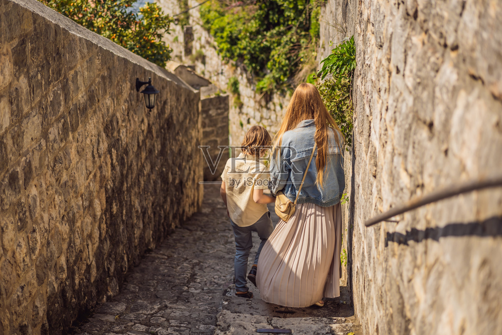 在一个阳光明媚的日子里，妈妈和儿子在黑山的佩拉斯特老城享受五彩缤纷的街道。黑山旅行的概念。在一个美丽的阳光明媚的日子里，蓝天白云，著名的科托尔湾的历史小镇佩拉斯特的全景景色照片摄影图片
