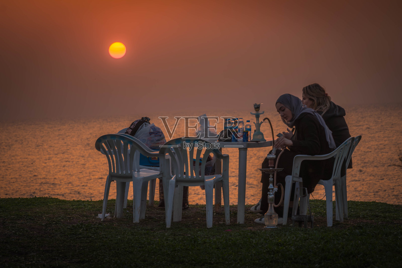 两个阿拉伯女孩朋友坐在地中海沿岸，抽着水烟，看着黎巴嫩泰尔(苏尔)的日落。照片摄影图片