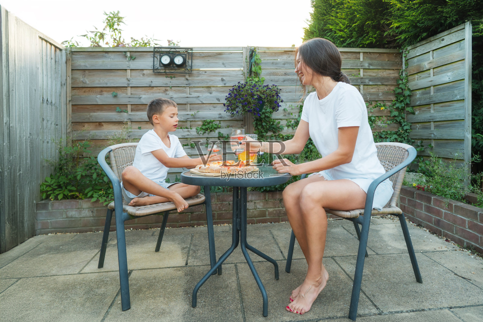 快乐的年轻母亲和儿子享受着新鲜空气的晚餐，坐在户外家具上吃着披萨和柠檬水。家庭团聚的快乐时光。活跃的童年。夏天在郊区的房子。照片摄影图片