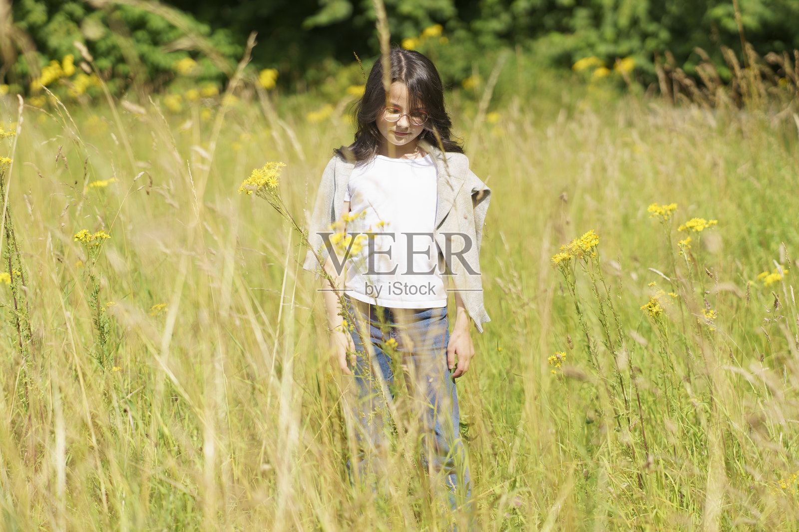 一个十几岁的小女孩戴着眼镜，穿着牛仔裤，t恤和一件毛衣披在肩上，站在齐腰深的天然草地上。小孩子的快乐在野花里。照片摄影图片