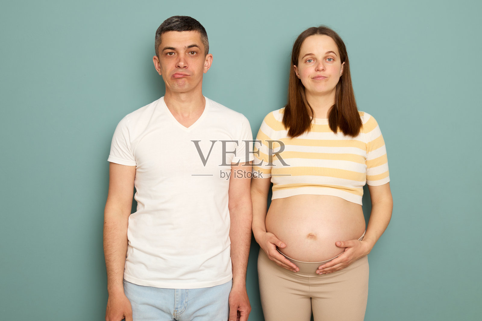 悲伤的男子和怀孕女子抚摸肚子，准父母面露悲伤，在绿松石背景下对着镜头照片摄影图片