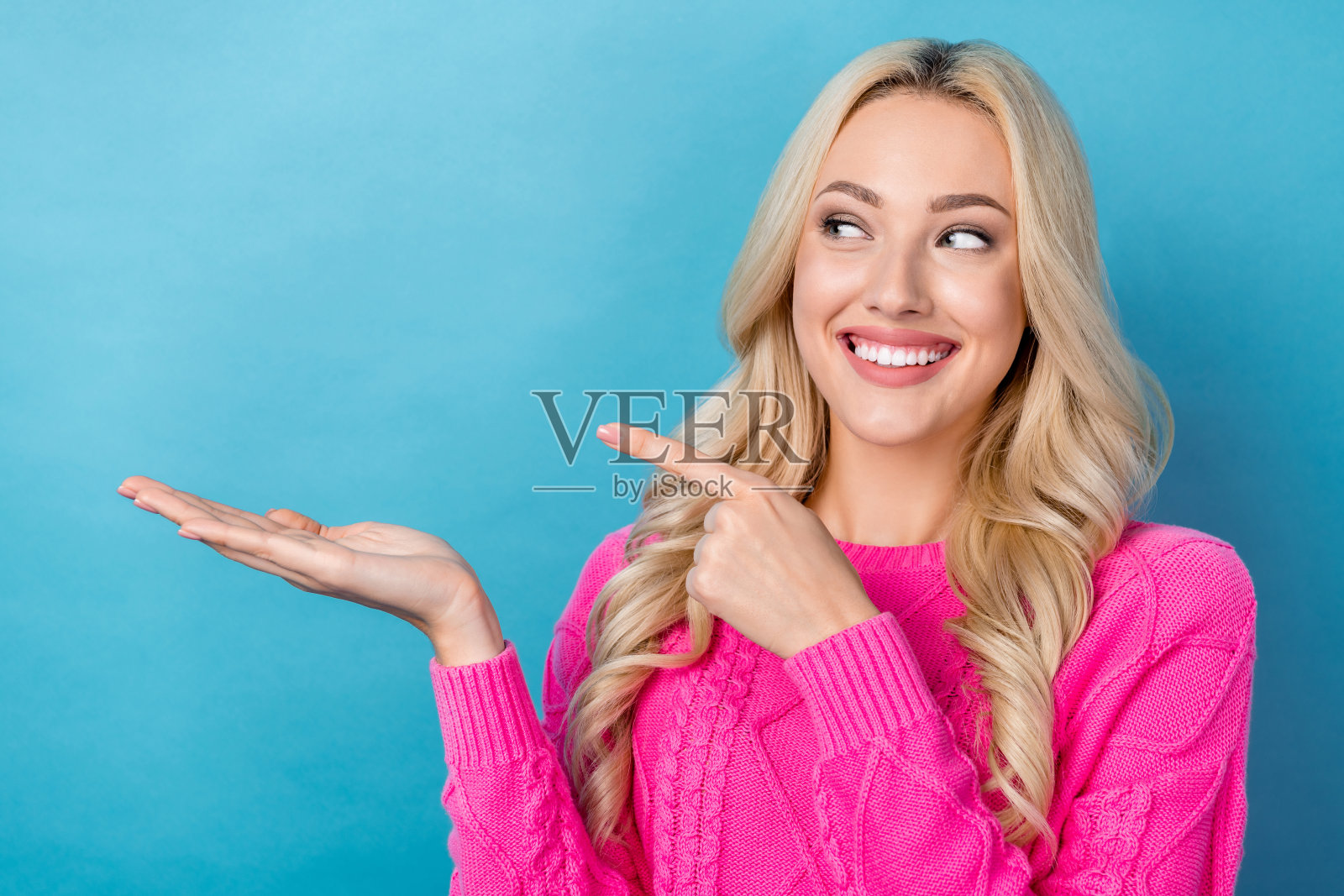 照片中，甜美开朗的女士穿着粉红色的毛衣，手指握着手臂，看着空无一物的蓝色背景照片摄影图片