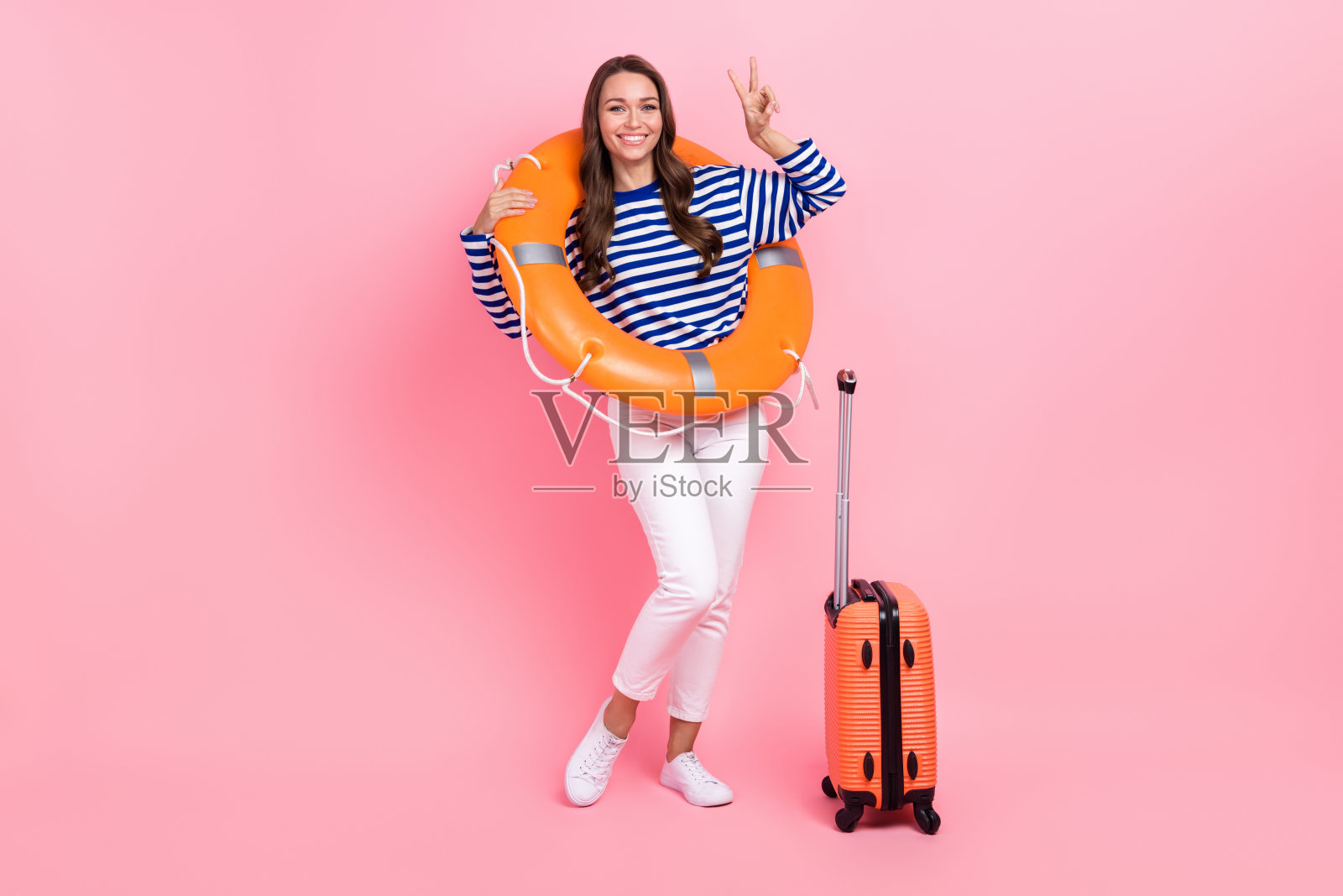 乐观女孩的全尺寸照片，穿着条纹衣服，白色裤子，肩上的救生圈，在粉红色的背景上显示v字照片摄影图片