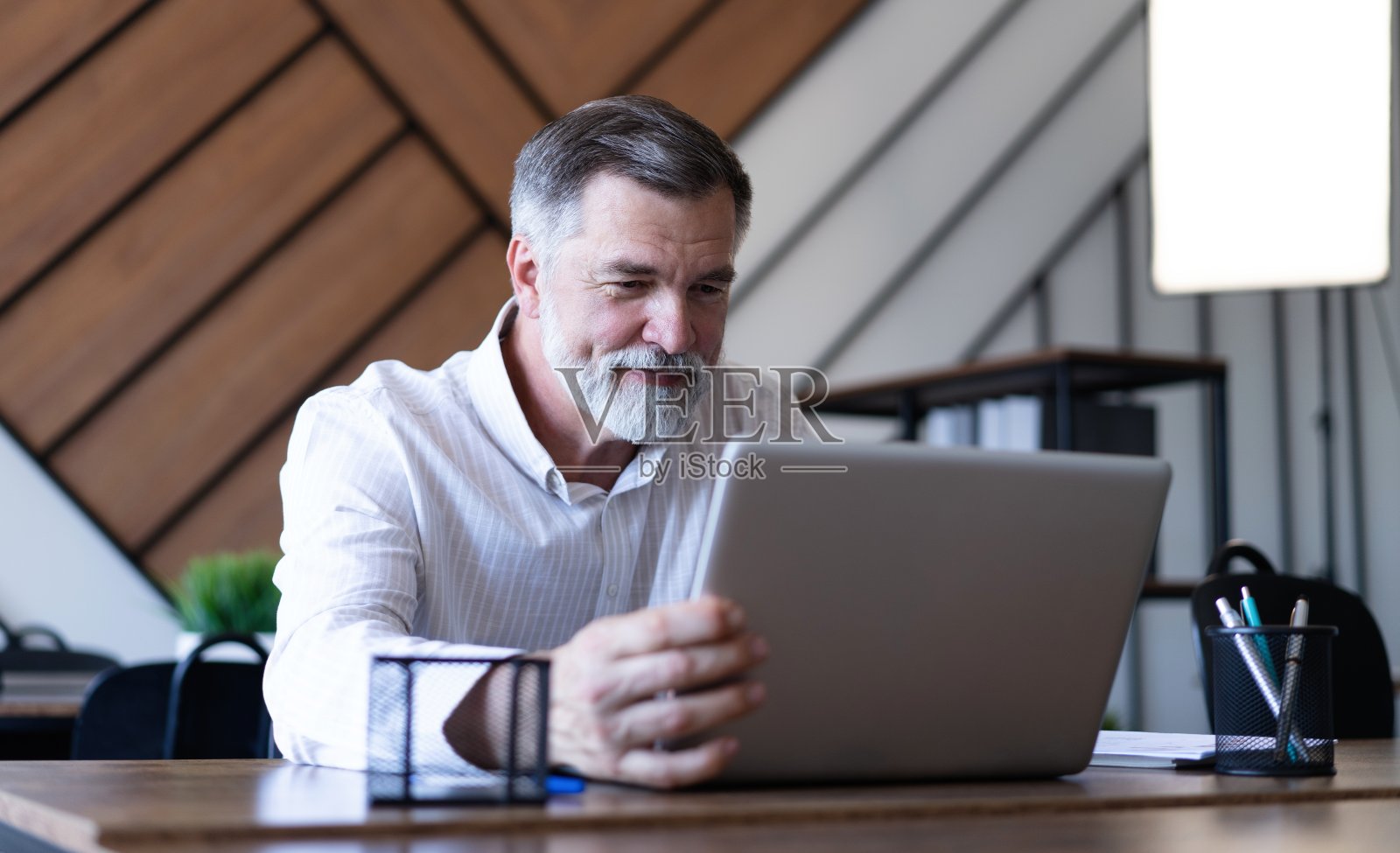 微笑的成熟的商人，穿着衬衫，坐在办公桌前使用笔记本电脑。快乐忙碌的职业中年商人投资者在电脑上工作，在办公室里望着远方照片摄影图片
