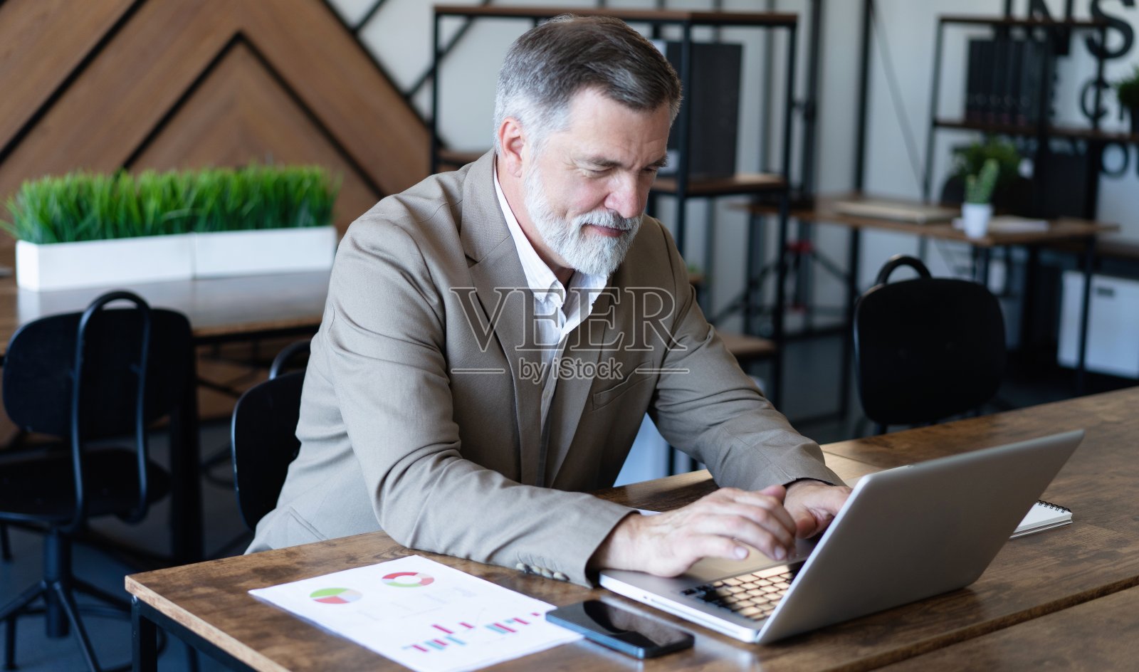 微笑的成熟的商人，穿着衬衫，坐在办公桌前使用笔记本电脑。快乐忙碌的职业中年商人投资者在电脑上工作，在办公室里望着远方照片摄影图片