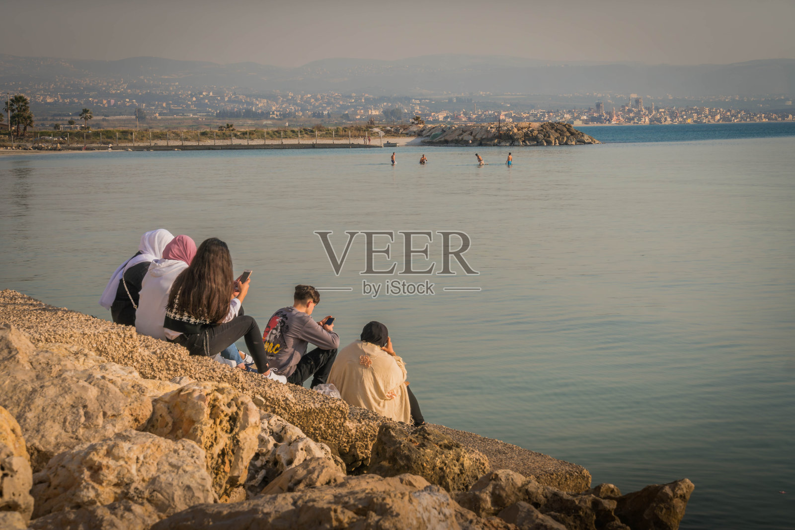 年轻的阿拉伯人在黎巴嫩地中海的岩石海岸与提尔(苏尔)城市的全景。照片摄影图片