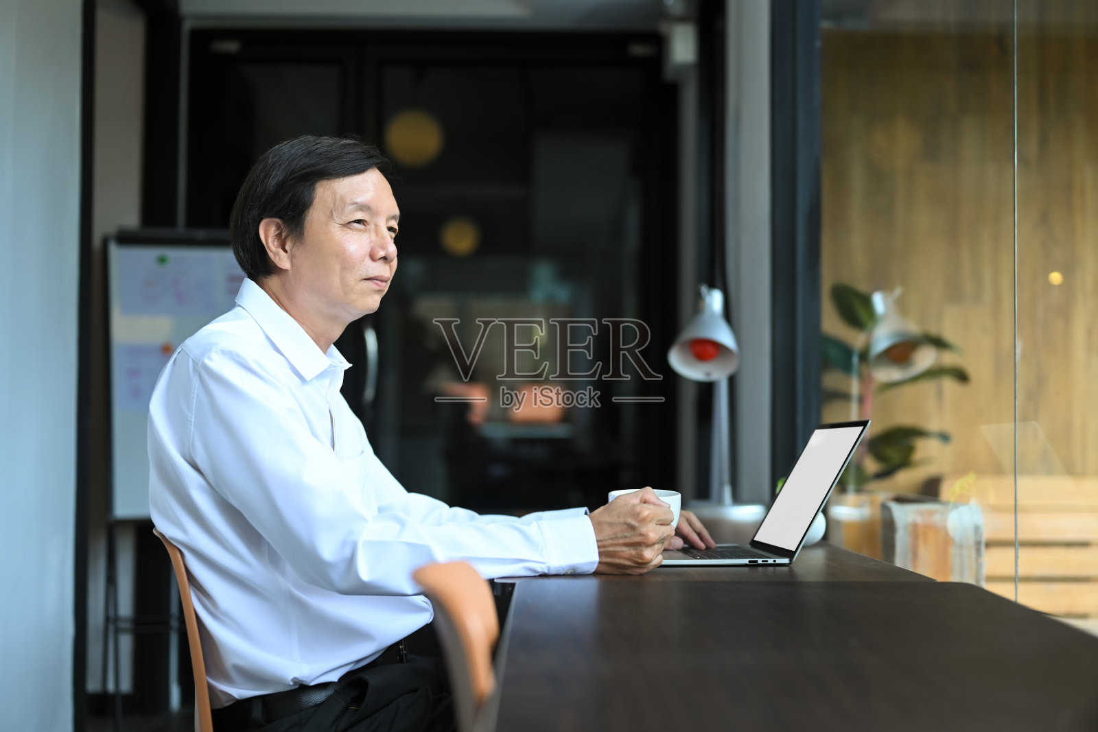 面带微笑的成熟商人，端着咖啡坐在办公桌前，目光移开，想着成功照片摄影图片