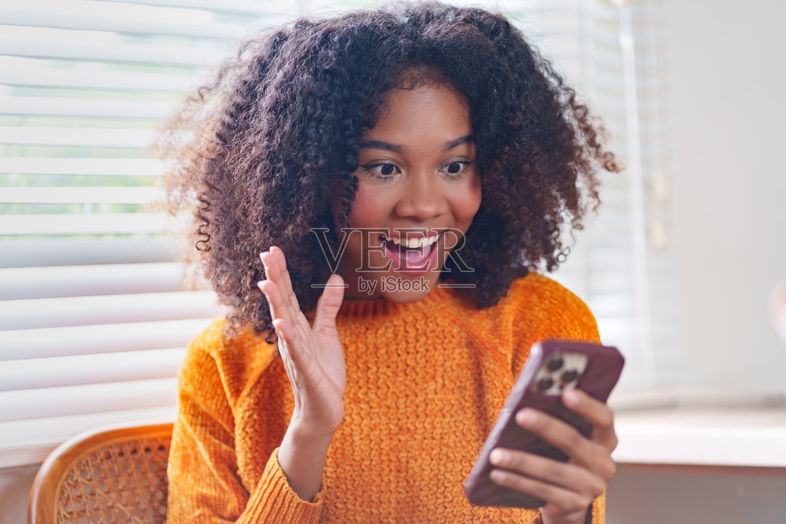 一名身穿橙色毛衣的非洲裔美国妇女在家里视频聊天时，兴奋地挥舞着智能手机。照片摄影图片