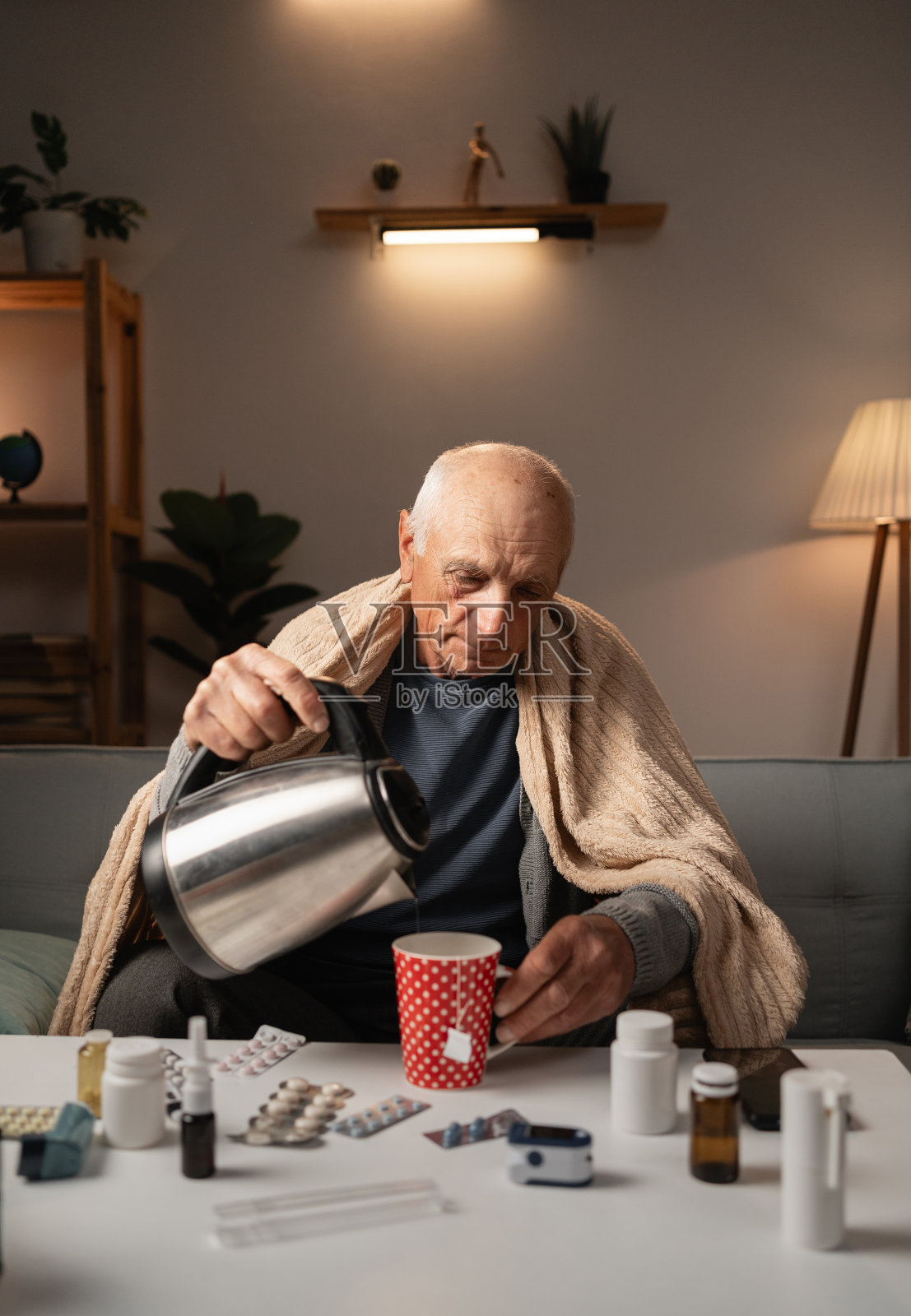 老病人坐在沙发上，从水壶里倒热水，泡茶，裹在毯子里，周围是药物照片摄影图片