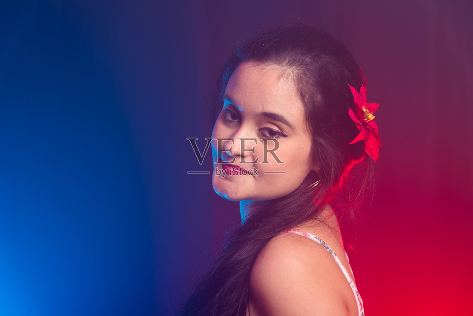 美丽的黑发女子穿着色彩鲜艳的裙子，左耳上戴着一朵红花，摆姿势拍照。照片摄影图片