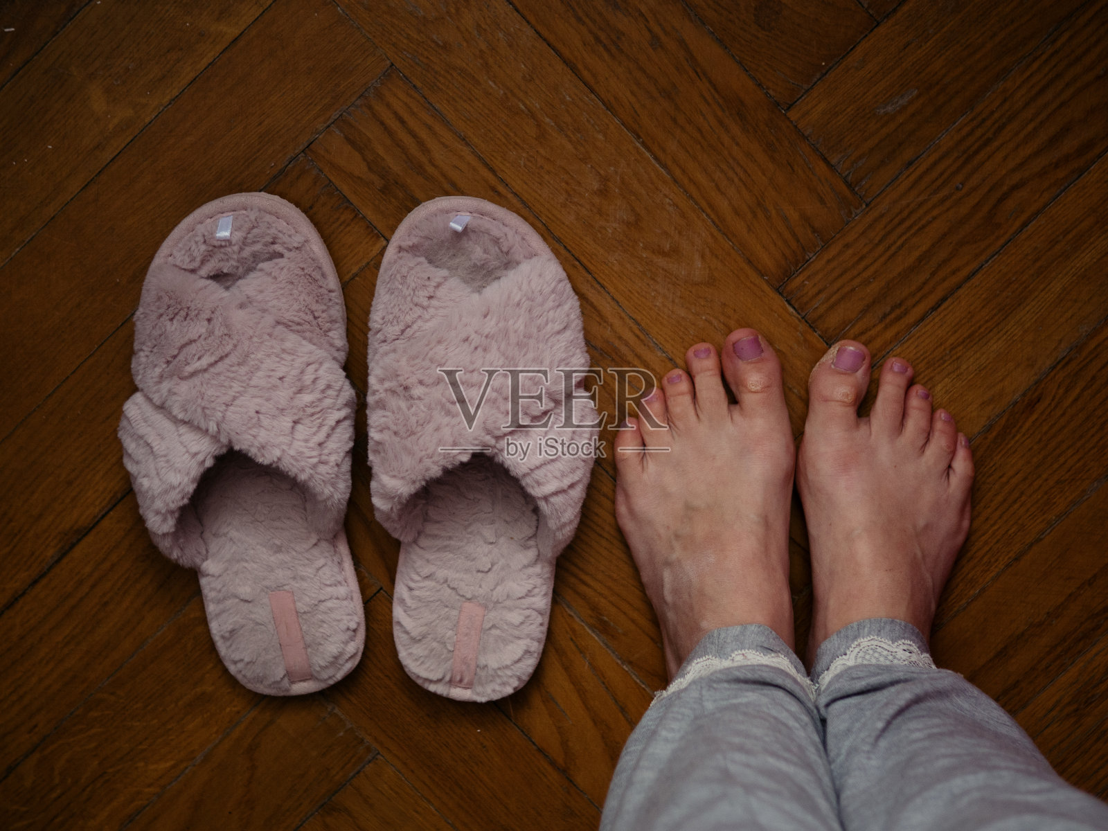 正上方的水平特写，一个穿着灰色睡衣的女人赤脚站在镶木地板上，旁边是粉红色的绒毛拖鞋，脚部有瑕疵。偷拍的家庭照片。照片摄影图片