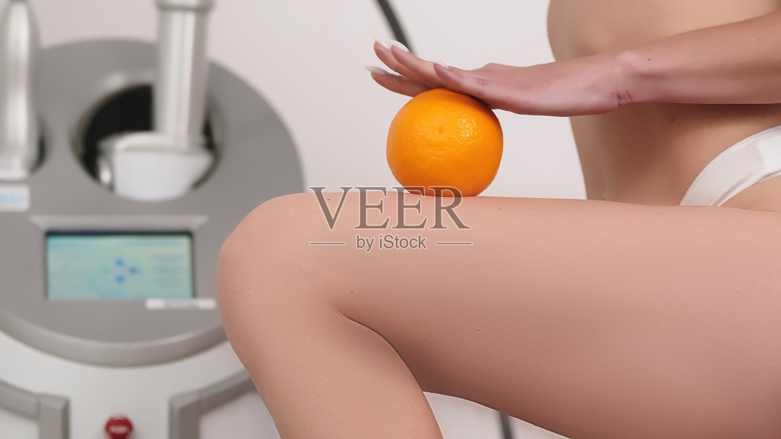 一名女子将一个橙子放在腿上，背景是一个按摩和提升装置。健康与美丽的理念。象征着对抗脂肪团和改善肤色。照片摄影图片