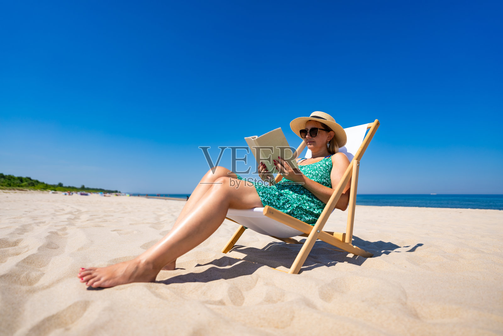 暑假在海滩上。在沙滩上晒日光浴和看书。美丽的女人穿着绿色的衣服，戴着米色的帽子，在阳光明媚的日子里坐在白色沙滩上的日光浴床上看书。在海滩上放松。前视图照片摄影图片
