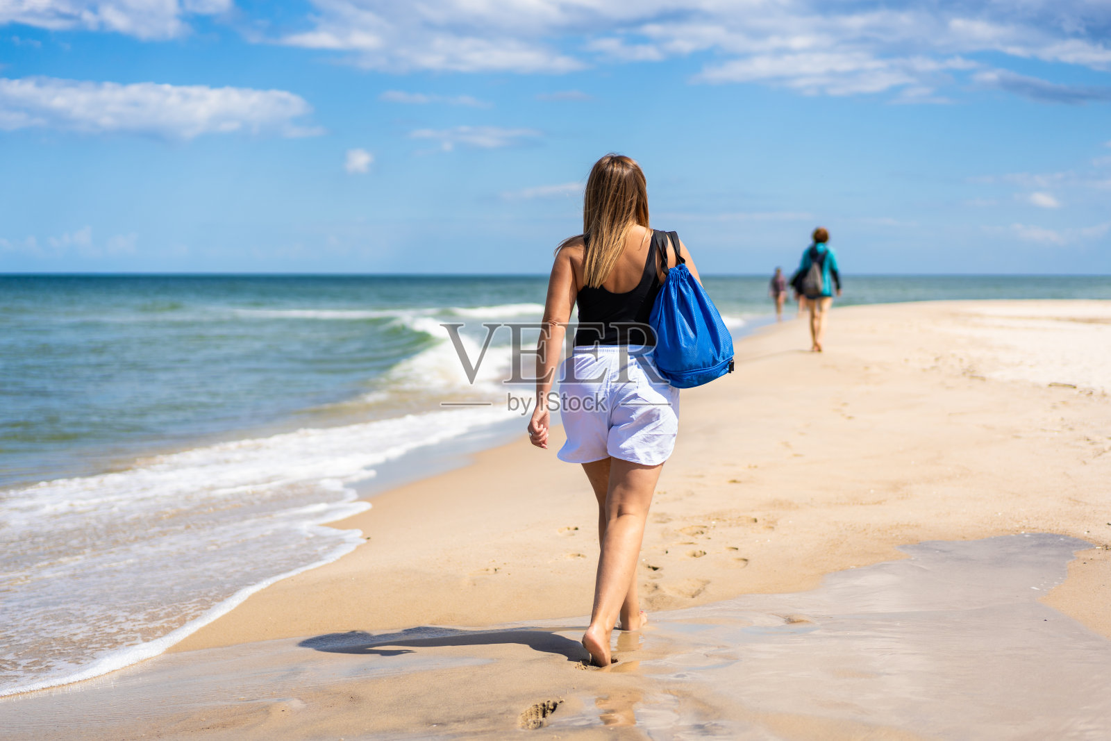 夏天在海滩上的体育活动。一个金色长发的女人，穿着黑色泳衣和白色运动短裤，戴着蓝色运动包的太阳镜，在阳光明媚的日子里走在沙滩上。照片摄影图片