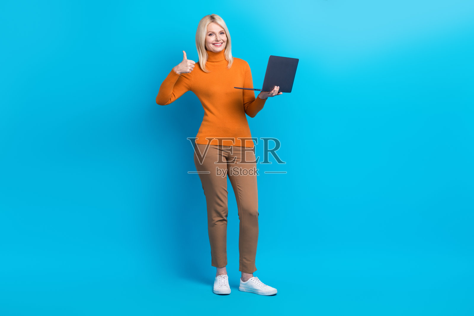 积极开朗的女性全身照片，穿着橙色运动衫，拿着笔记本电脑，拇指向上，孤立在蓝色背景上照片摄影图片