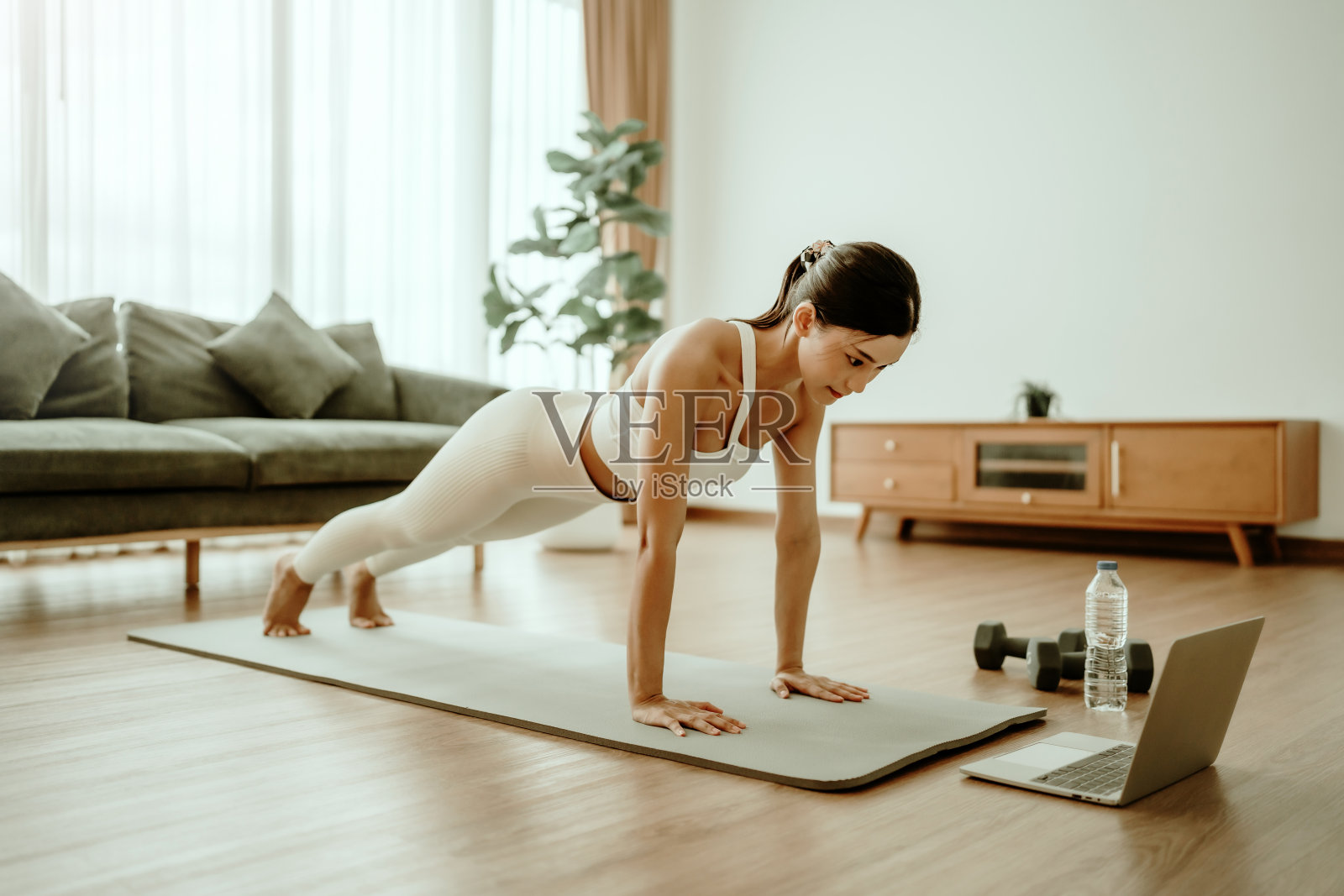 年轻的亚洲女性穿着运动服在训练垫上做平板支撑，同时在客厅的笔记本电脑上观看在线锻炼教程。运动及康乐概念照片摄影图片