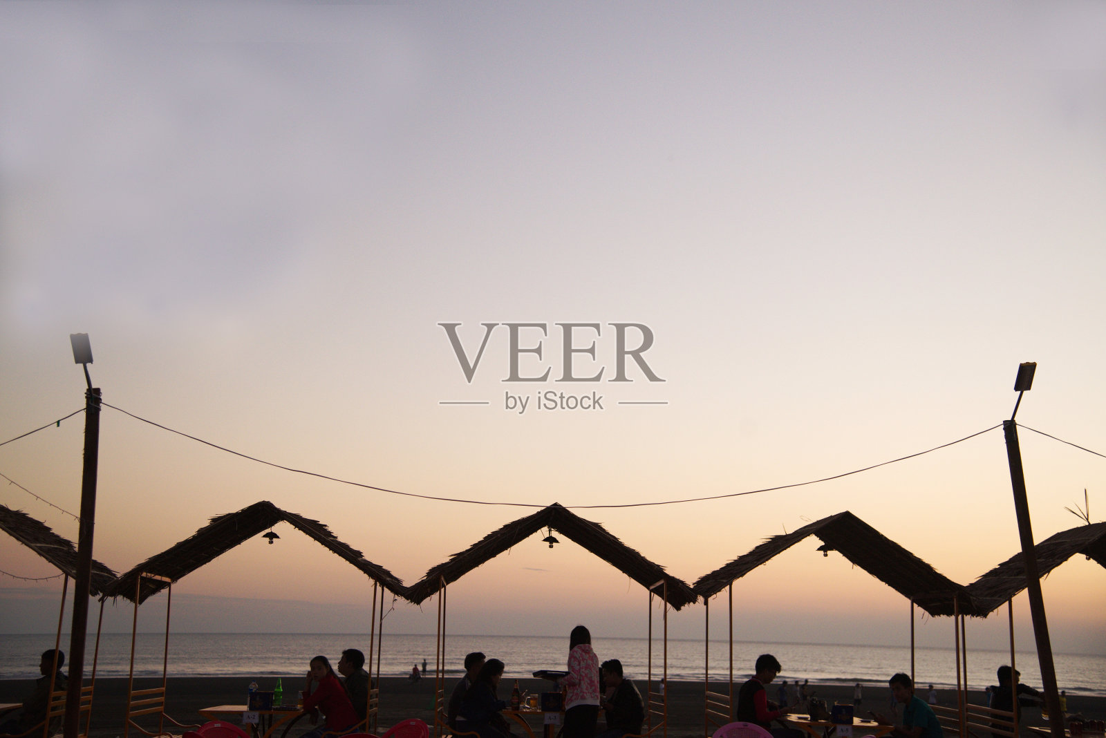 日落时分，实兑海滩上，身份不明的游客剪影坐在稻草屋顶的桌子旁吃饭。照片摄影图片