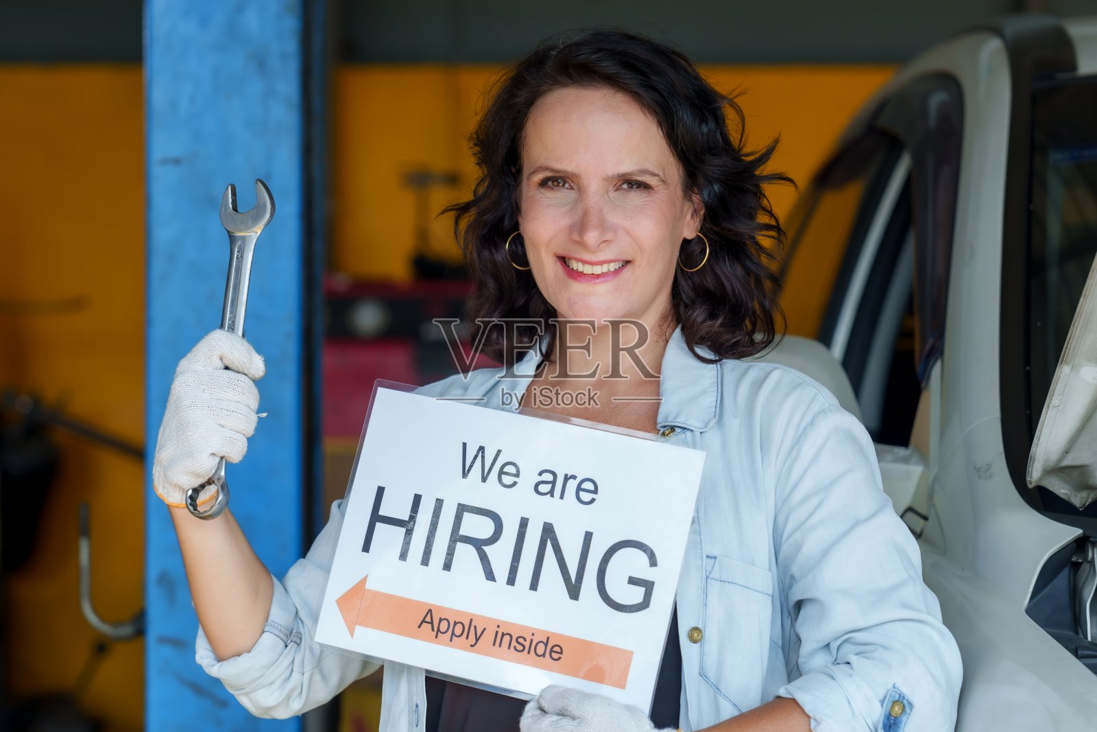 白人女性成年机械师拿着扳手和招聘标志在车库里微笑。白色的手套。以自信和友好的态度促进汽车维修行业的工作机会。照片摄影图片