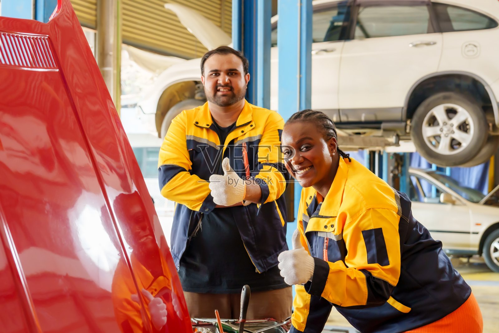 男技工和女技工在车库里一辆红色凸起的引擎盖前竖起大拇指。穿着黄色和蓝色的制服。体现团队合作，认可，信心完成汽车维修任务。照片摄影图片