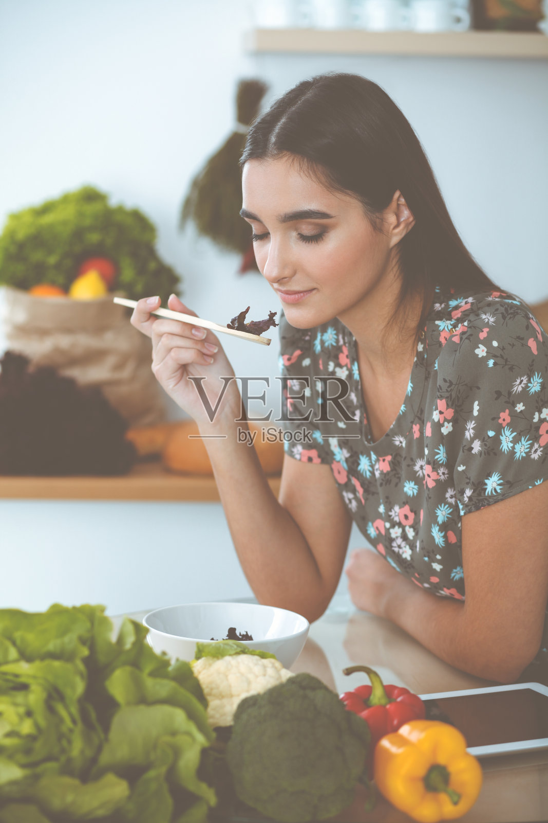 一位迷人的黑发年轻女子坐在厨房的餐桌旁，品尝着一种美味沙拉的新配方。平板电脑是最好的食谱照片摄影图片
