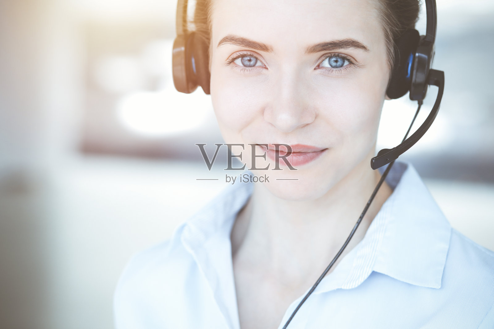 在阳光明媚的呼叫中心办公室里，戴着耳机的美丽女专家在线帮助客户，迷人地微笑着。商务人士概念照片摄影图片