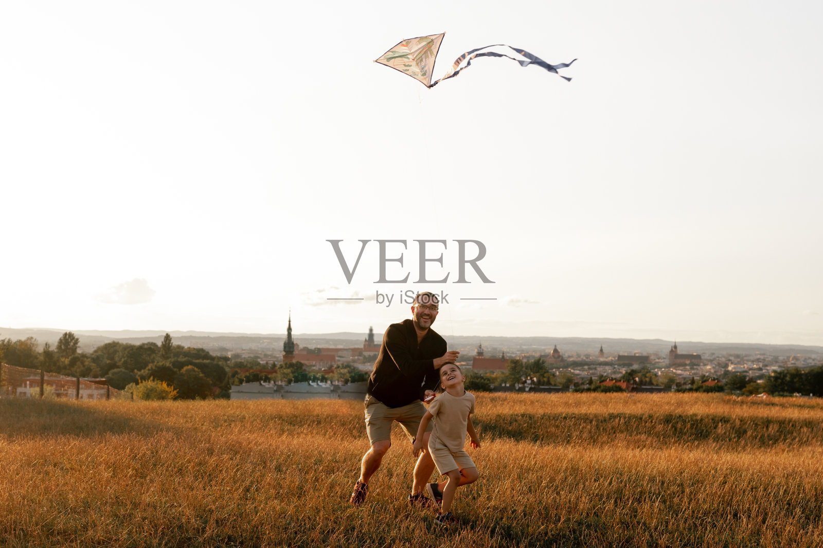 快乐的父亲和儿子在草地上玩风筝，日落，夏日。有趣的家庭时光。快乐的小孩和爸爸一起放风筝。照片摄影图片