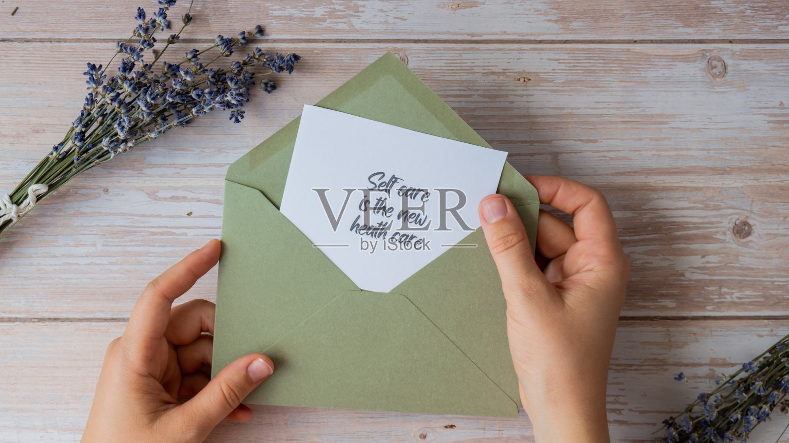 女性的手从绿色信封里拿着纸卡片，上面写着“自我保健是新的健康保健”。薰衣草的花。俯视图，平铺。心理、精神健康、自我照顾、幸福的概念照片摄影图片