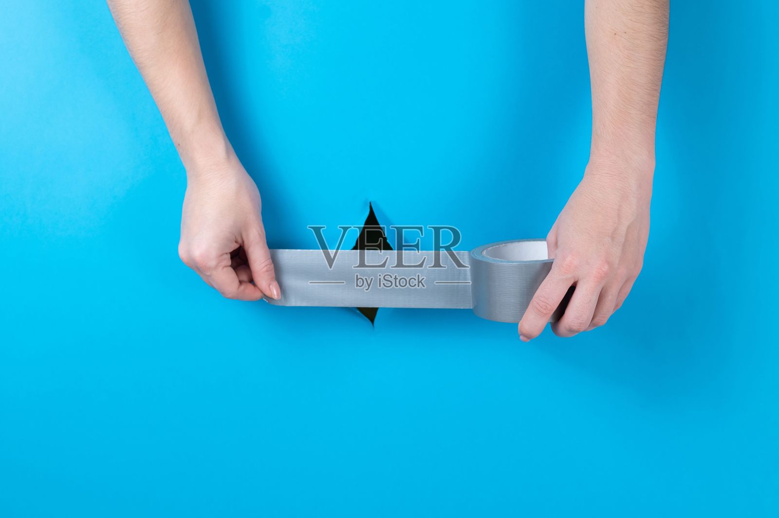 一个女人把银色胶带粘在蓝色背景上的洞上。照片摄影图片