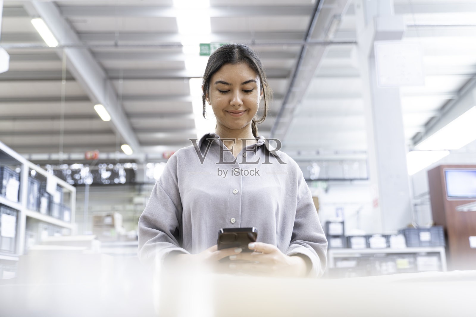 穿着蓝色工作服，斜着身子工作的女商人，一边说话，一边看短信，一边在工厂里用智能手机发短信和社交媒体平台照片摄影图片