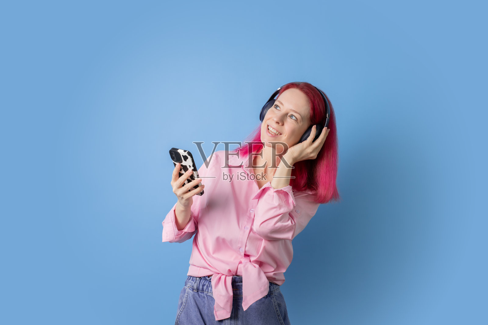 粉色头发的女人拿着蓝色背景的手机和耳机跳舞，在智能手机上享受音乐，时尚而无忧无虑，快乐地微笑着。照片摄影图片