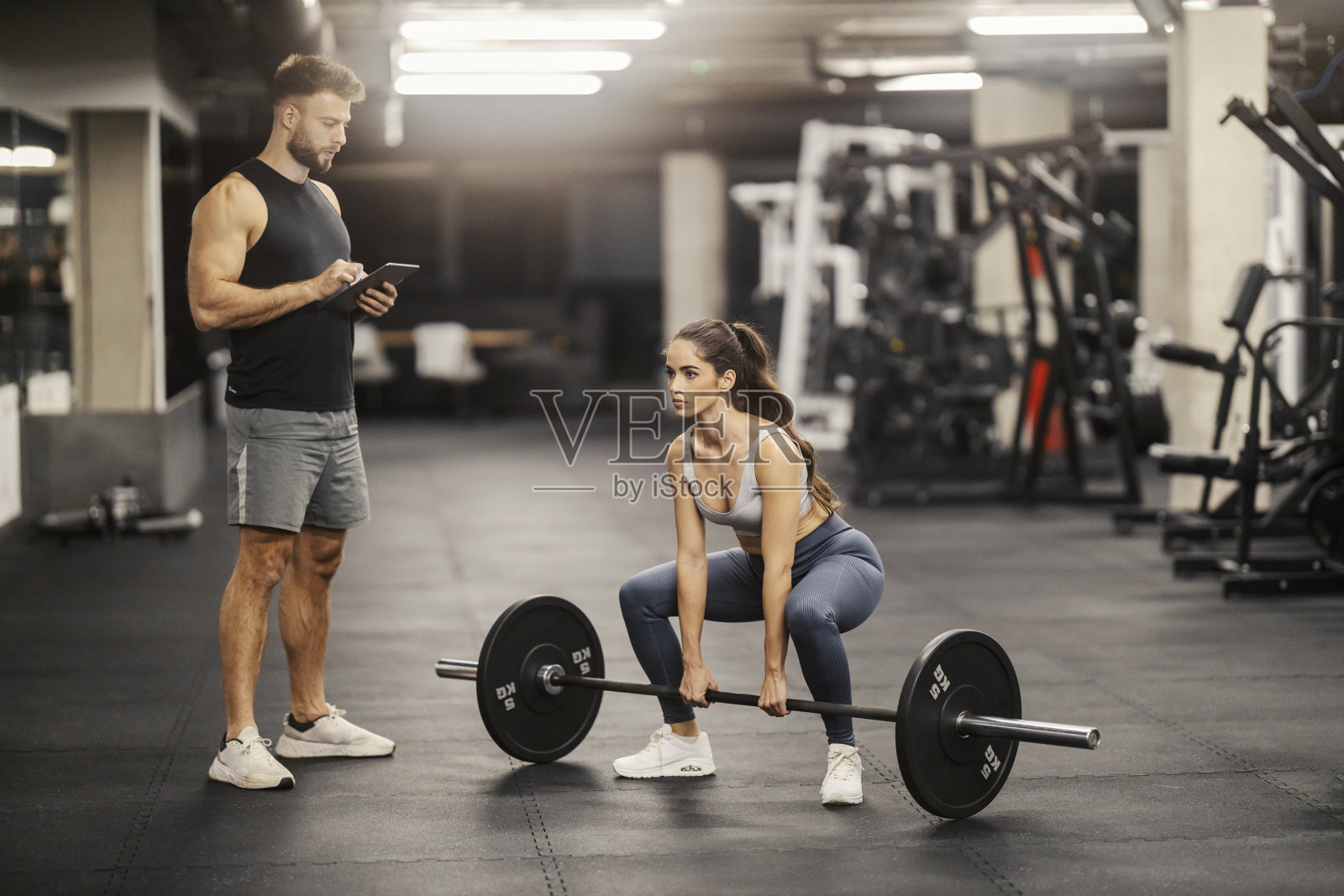 一名女运动员在健身房里蹲着举杠铃，她的私人教练正在看着她，并在平板电脑上记录她的进步。照片摄影图片