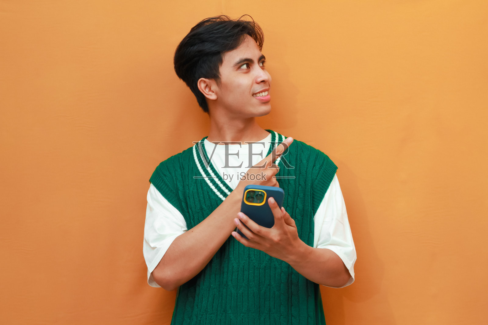 快乐的年轻亚洲人穿着绿色毛衣，拿着手机，用手指着照片摄影图片