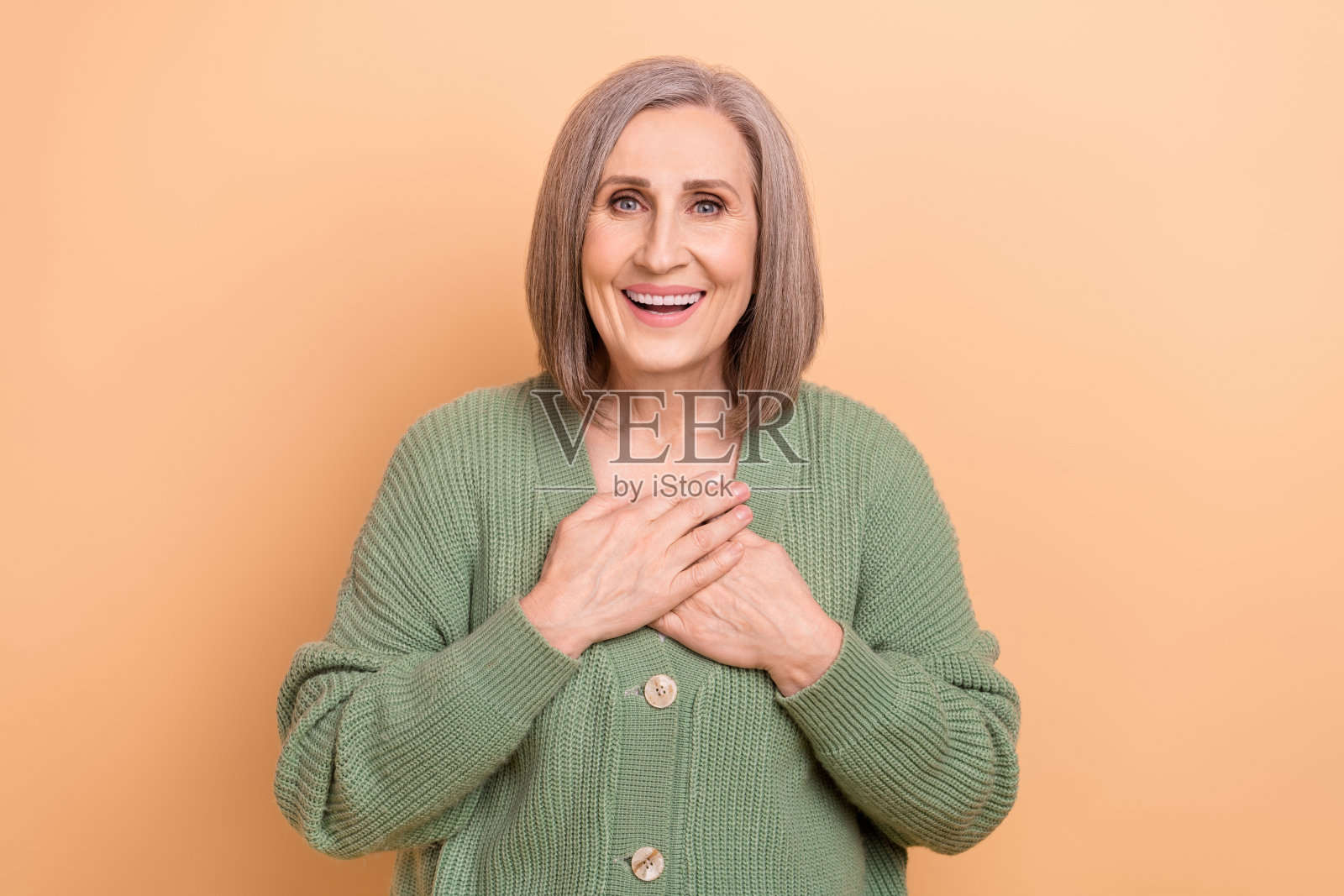 照片愉快的退休妇女与灰色的头发，穿着卡其开襟羊毛衫手在胸前孤立的米色背景照片摄影图片