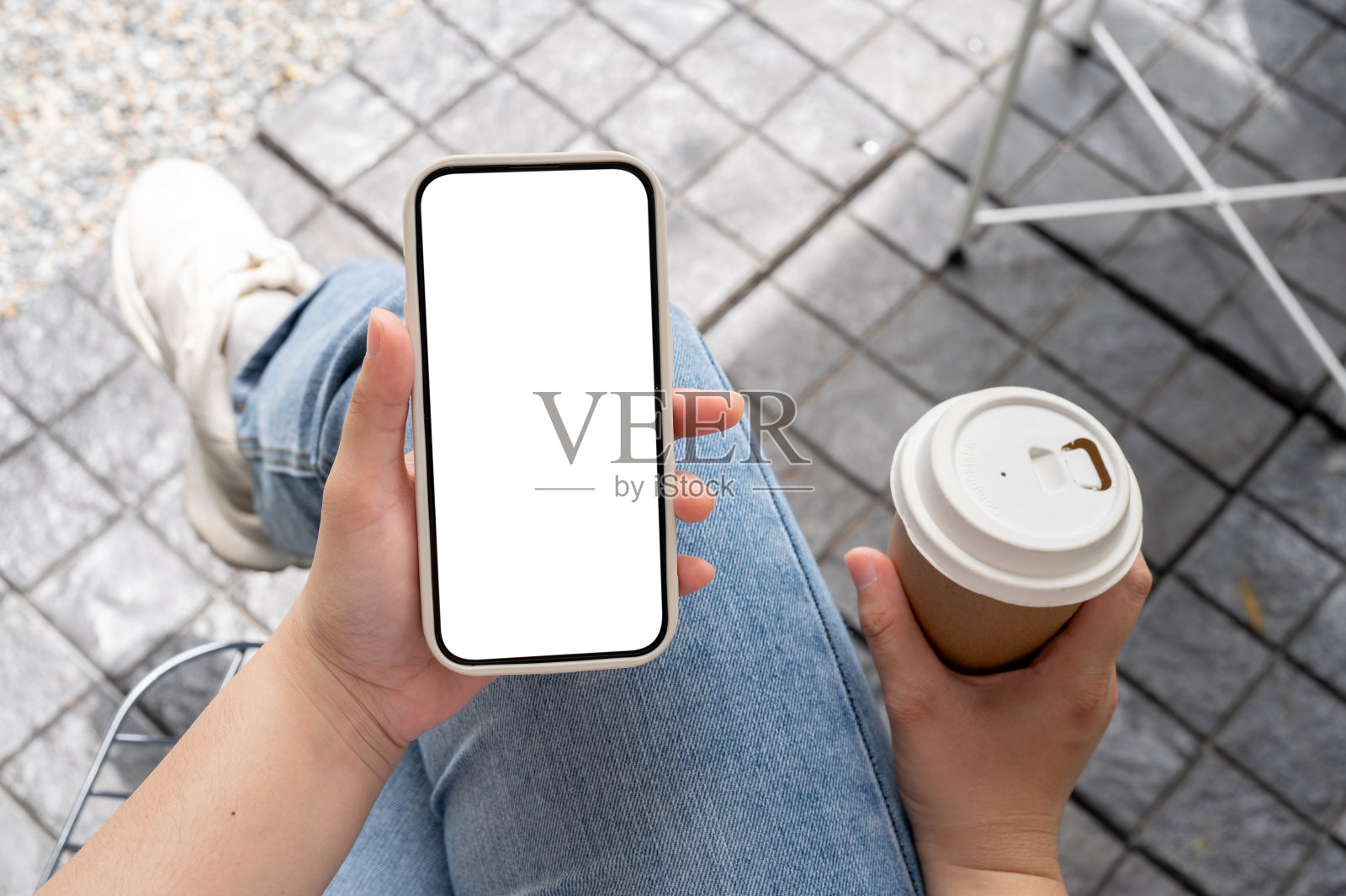 一个穿着牛仔裤的女人坐在户外，手里拿着咖啡杯，用着她的智能手机。照片摄影图片