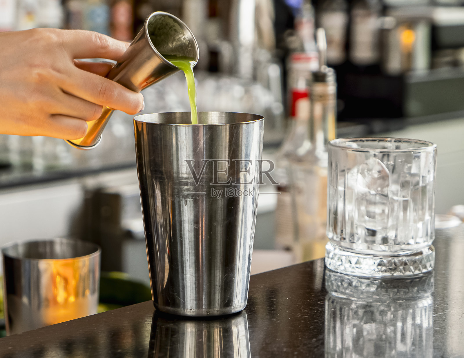 用手把绿色的液体从一个小金属壶里倒进吧台上一个更大的摇酒器里，旁边放着一个空杯子照片摄影图片