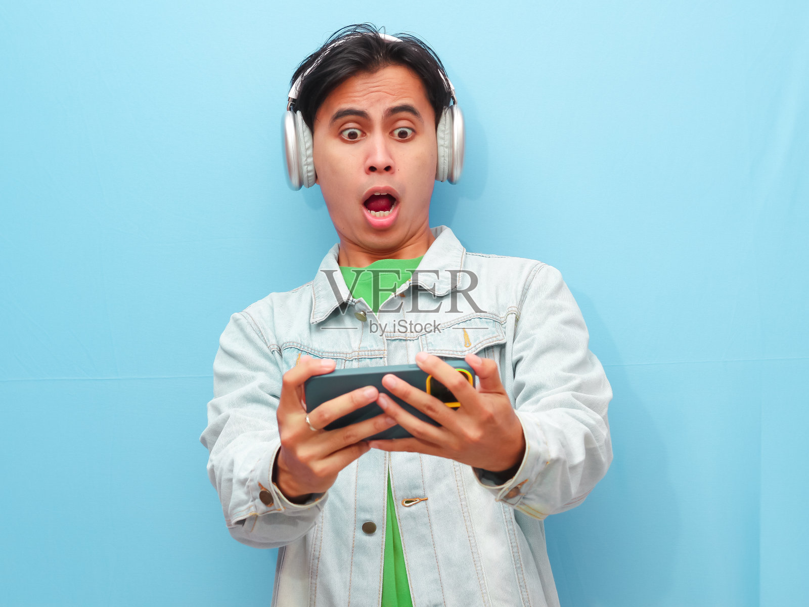 兴奋快乐的年轻亚洲男性玩家使用智能手机玩游戏，在移动视频游戏应用程序中观看数字视频流，手持手机孤立在蓝色背景下。照片摄影图片