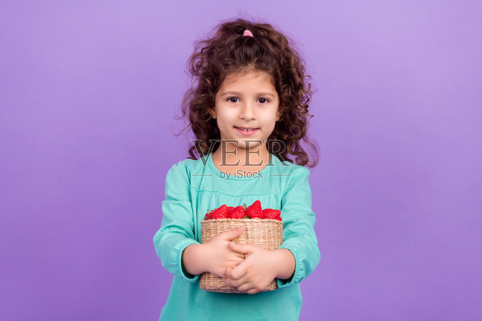 照片中可爱可爱的小孩穿着蓝绿色的衬衫，收集满草莓篮子，孤立的紫罗兰色背景照片摄影图片
