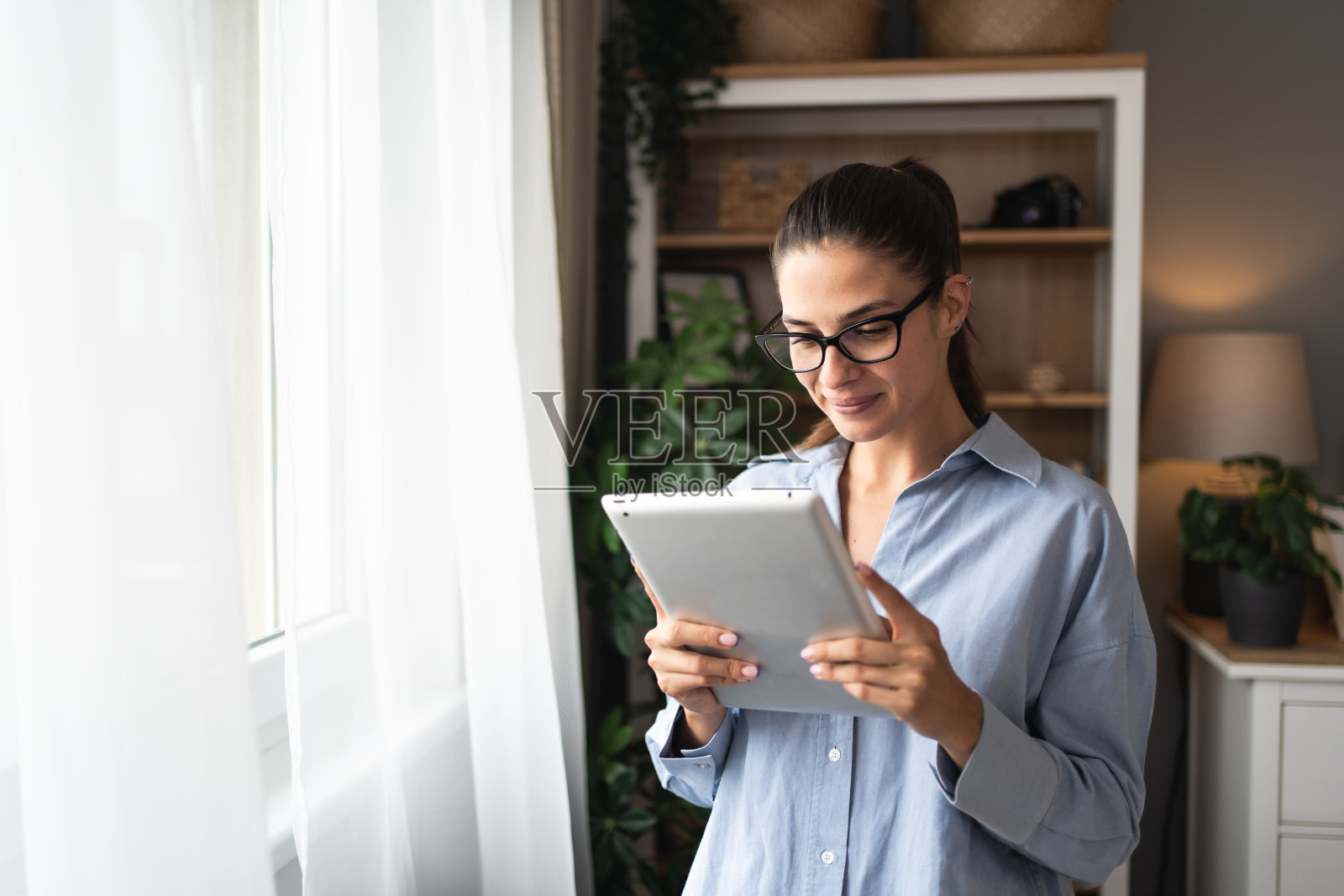 房地产经纪人用平板电脑向窗外看。年轻的女商人在家工作，使用电子设备。成功的专业职业平面设计师女性在工作之余休息。照片摄影图片