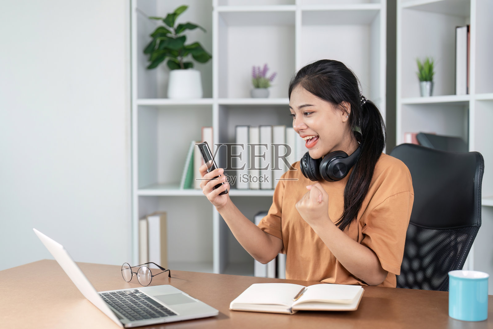 快乐的年轻女子庆祝令人兴奋的消息，而看着她的手机在现代家庭办公环境照片摄影图片